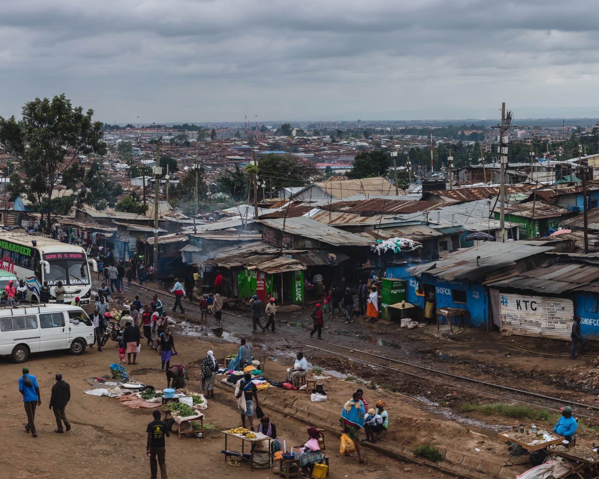 Vendors and pedestrians along a railway-track market in Nairobi's Kibera settlement, with a bus among closely packed shops